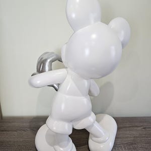 May include: A white Mickey Mouse figurine with a silver-colored hand. The figurine is in a walking pose, with a glossy finish. The sculpture is a decorative item.