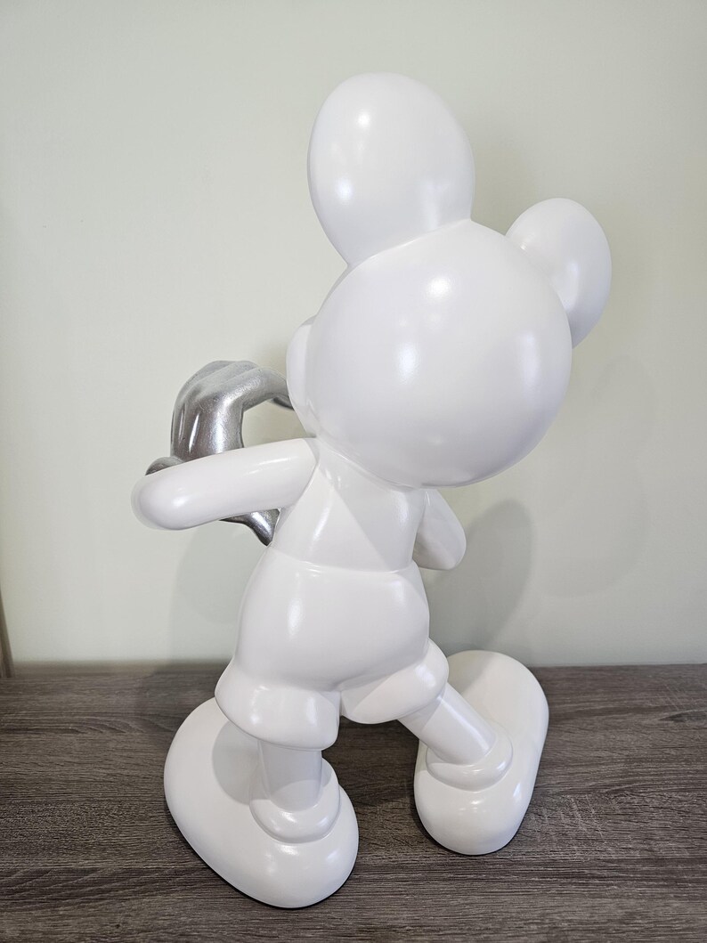 May include: A white Mickey Mouse figurine with a silver-colored hand. The figurine is in a walking pose, with a glossy finish. The sculpture is a decorative item.