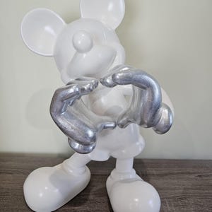 May include: A white Mickey Mouse figurine with silver-colored hands forming a heart shape. The figurine has large, rounded ears and feet, and is posed in a standing position. The background is a neutral color.