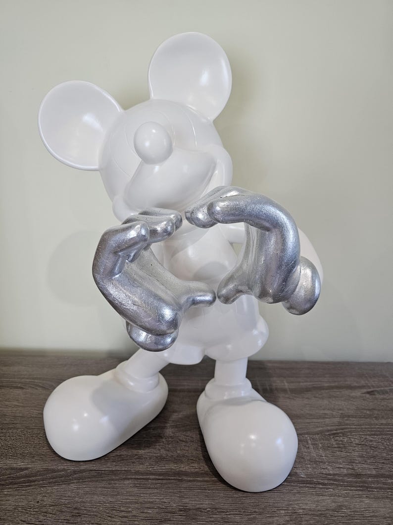 May include: A white Mickey Mouse figurine with silver-colored hands forming a heart shape. The figurine has large, rounded ears and feet, and is posed in a standing position. The background is a neutral color.