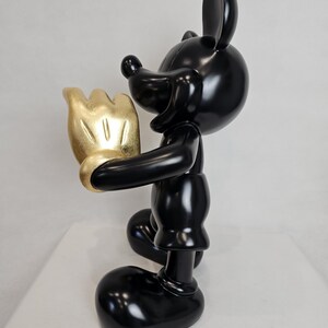 May include: A glossy black figurine of a cartoon mouse holding a gold-colored hand. The mouse has large ears, a round head, and a classic design. The figurine is on a white surface.