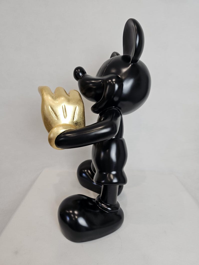 May include: A glossy black figurine of a cartoon mouse holding a gold-colored hand. The mouse has large ears, a round head, and a classic design. The figurine is on a white surface.