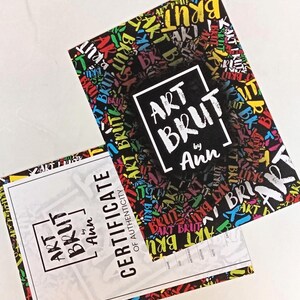 May include: A certificate of authenticity for art brut by Ann. The certificate is white with black text and a black and white logo with the words "Art Brut by Ann". The background of the certificate is a colorful pattern of the words "Art Brut" in various colors.