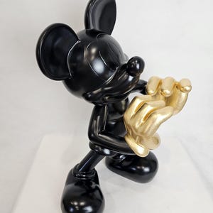 May include: A glossy black Mickey Mouse figurine with gold-colored hands. The sculpture is posed in a dynamic stance, with one hand raised and the other near the face. The figurine is on a white surface.
