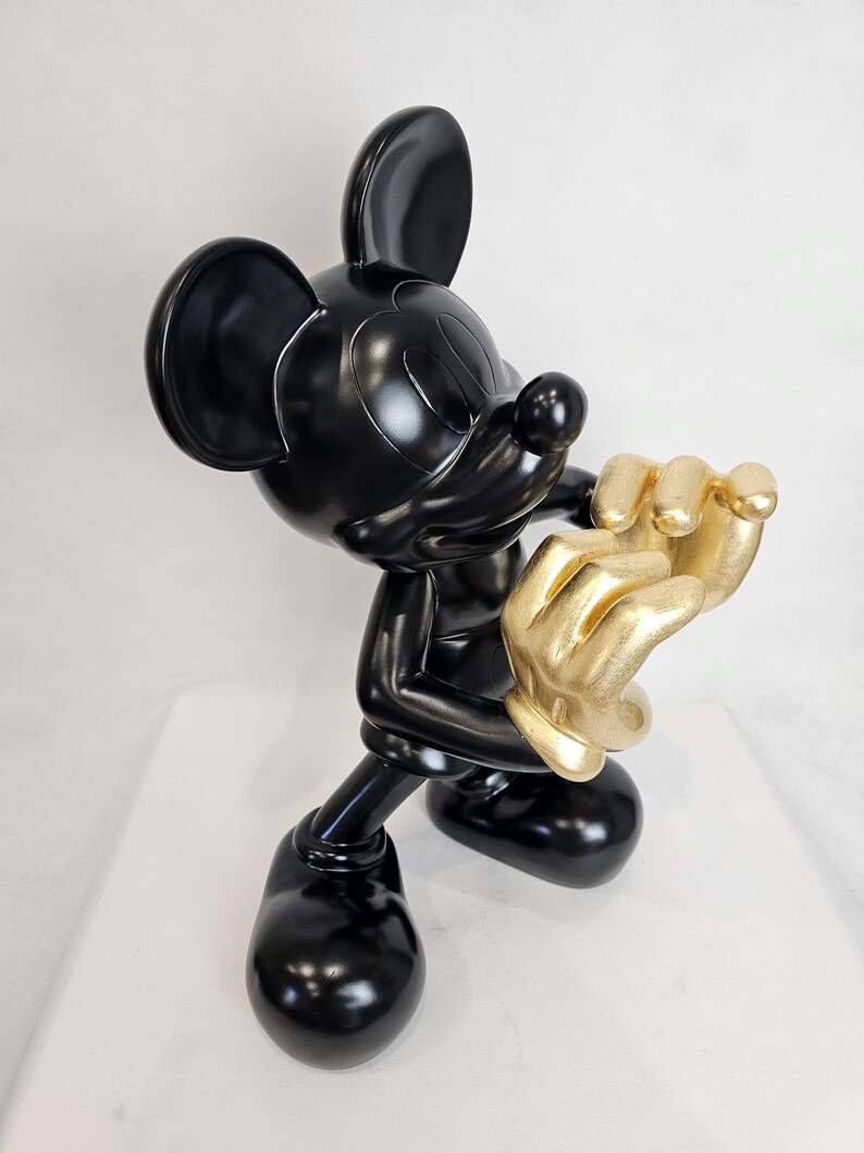 May include: A glossy black Mickey Mouse figurine with gold-colored hands. The sculpture is posed in a dynamic stance, with one hand raised and the other near the face. The figurine is on a white surface.