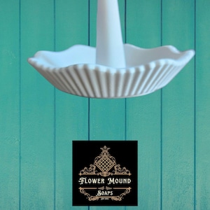 May include: A white ceramic ring holder with a fluted, scalloped dish and a central cone-shaped post. The holder is set against a teal-colored wooden background. The logo for Flower Mound Soaps is in the lower portion of the image.