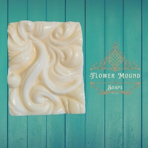 May include: Ivory-colored soap bar with a swirling, embossed design. The soap is rectangular and sits against a teal wooden background. The text "Flower Mound Soaps" is visible in the image.