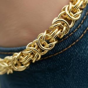 May include: Close-up of a gold-coloured chain bracelet with a Byzantine weave design. The bracelet features interlocking, rounded links. The bracelet is set against a denim background.