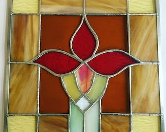 Op de afbeelding: Een glas-in-loodraam met een gestileerde rode bloem met gele en groene accenten. Het raam is ingelijst met zilver.