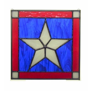 May include: A square stained glass panel featuring a white star on a blue background, framed by a red border. The panel has a hanging loop at the top. The design evokes patriotic themes.