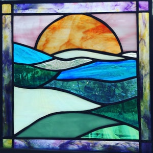 Puede incluir: Una ventana de vitrales que representa una puesta de sol sobre un paisaje con agua azul y colinas verdes. El sol es de un naranja y amarillo brillantes, y el cielo es de un rosa suave.