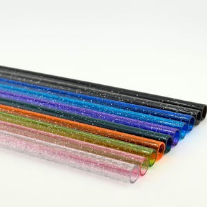 May include: A collection of colorful, glitter-filled straws. The straws are arranged in a diagonal row, showcasing a variety of colors including pink, green, orange, blue, and black. Each straw has a clear, cylindrical shape with sparkling glitter.