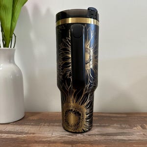 Puede incluir: Vaso de viaje negro con un dise&ntilde;o de girasol dorado. El vaso tiene un asa negra y un borde dorado. El dise&ntilde;o del girasol est&aacute; grabado en la superficie negra. El vaso est&aacute; sobre una superficie de madera.