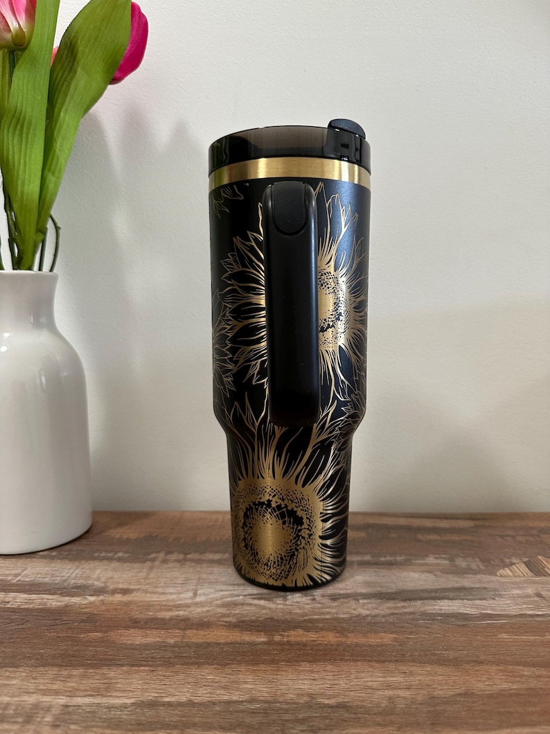 Puede incluir: Vaso de viaje negro con un dise&ntilde;o de girasol dorado. El vaso tiene un asa negra y un borde dorado. El dise&ntilde;o del girasol est&aacute; grabado en la superficie negra. El vaso est&aacute; sobre una superficie de madera.