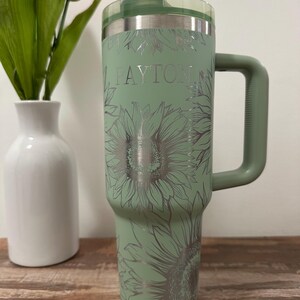 Puede incluir: Un vaso t&eacute;rmico verde salvia con asa, con un dise&ntilde;o de girasoles y el nombre "PAYTON" en plata. El vaso tiene una tapa transparente y est&aacute; dise&ntilde;ado para bebidas.