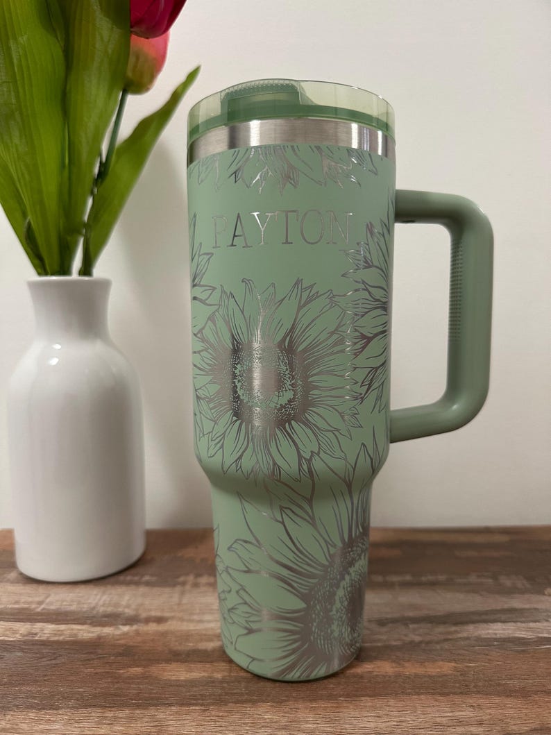 Puede incluir: Un vaso t&eacute;rmico verde salvia con asa, con un dise&ntilde;o de girasoles y el nombre "PAYTON" en plata. El vaso tiene una tapa transparente y est&aacute; dise&ntilde;ado para bebidas.