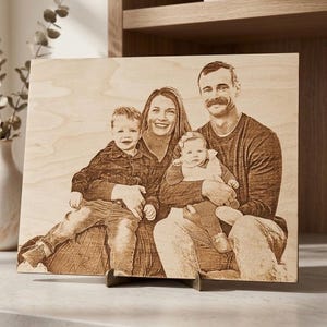 May include: A wooden family portrait, laser-etched with a photograph of a family of four. The image is displayed on a wooden plaque, supported by a black stand. The family includes two parents and two young children.