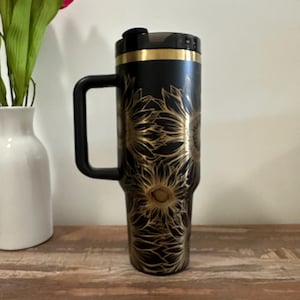 Puede incluir: Taza de viaje negra con un dise&ntilde;o de girasol dorado. La taza tiene un asa y una tapa negras con una banda dorada. La taza est&aacute; sobre una superficie de madera.