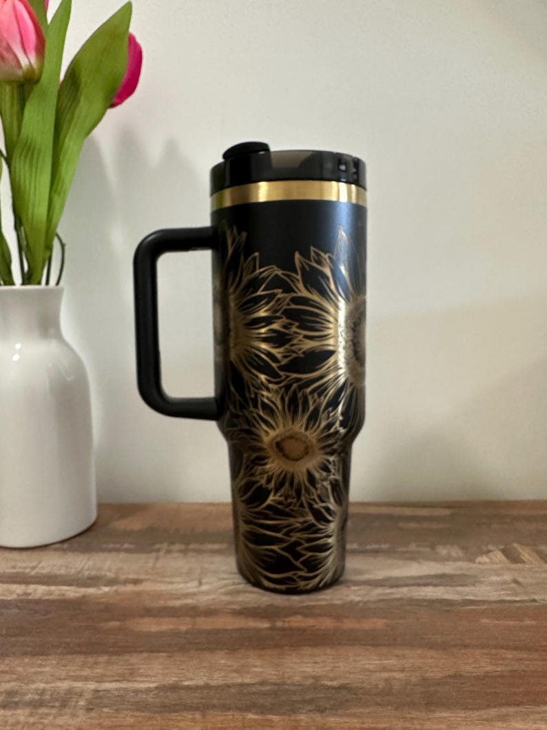 Puede incluir: Taza de viaje negra con un dise&ntilde;o de girasol dorado. La taza tiene un asa y una tapa negras con una banda dorada. La taza est&aacute; sobre una superficie de madera.