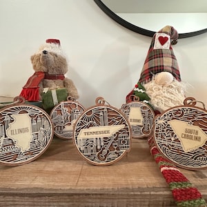 May include: Wooden state-shaped ornaments with laser-cut designs. The ornaments feature the names of states like Illinois and Tennessee, along with iconic imagery. A teddy bear and a gnome are in the background.