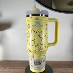 Puede incluir: Vaso Stanley Quencher H2.0 amarillo con asa y pajita. El vaso está decorado con ilustraciones de temática dental y el nombre "Paige". Tiene una capacidad de 1,18 litros y está diseñado para la hidratación durante todo el día.