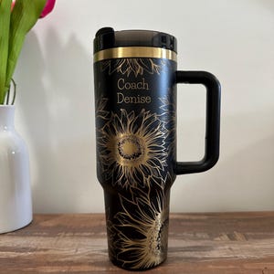 Puede incluir: Taza de viaje negra con un dise&ntilde;o de girasol dorado y el texto "Coach Denise". La taza tiene un asa y una tapa negras. El dise&ntilde;o presenta girasoles dorados detallados sobre un fondo negro.