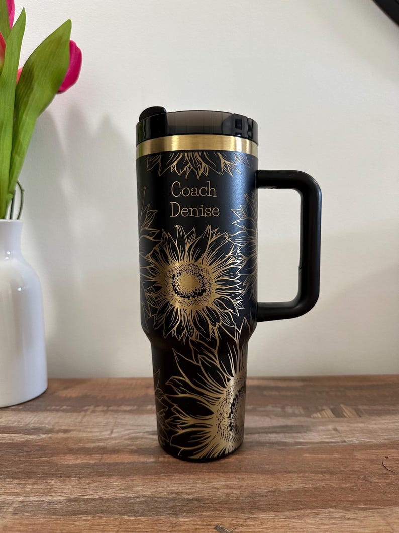 Puede incluir: Taza de viaje negra con un dise&ntilde;o de girasol dorado y el texto "Coach Denise". La taza tiene un asa y una tapa negras. El dise&ntilde;o presenta girasoles dorados detallados sobre un fondo negro.