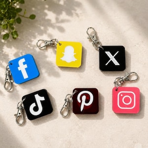 May include: A collection of six square keychains featuring social media logos including Facebook, Snapchat, X, TikTok, Pinterest, and Instagram. Each keychain is made of coloured plastic with a white logo and includes a silver metal lobster clasp.