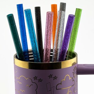 May include: A collection of colorful glitter straws in a purple mug. The straws are various colors, including green, blue, pink, and orange. The mug has a gold rim and a handle. The mug is decorated with gold cloud, star, and lightning bolt designs.