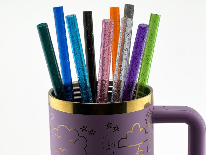 May include: A collection of colorful glitter straws in a purple mug. The straws are various colors, including green, blue, pink, and orange. The mug has a gold rim and a handle. The mug is decorated with gold cloud, star, and lightning bolt designs.