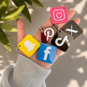 May include: A collection of square social media keychains featuring the logos for Instagram in pink, Pinterest in brown, X in black, TikTok in black, Snapchat in yellow, and Facebook in blue. Each keychain is attached to a small metal ring.