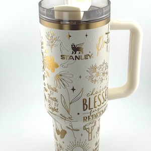May include: Cream Stanley travel mug with a clear lid and straw. It has gold floral and star patterns, plus inspirational text. A large handle is attached. The Stanley logo is visible on the side.