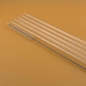 Peut inclure: Ensemble de pailles en verre transparentes avec une brosse de nettoyage. Les pailles sont cylindriques et légèrement courbées. La brosse de nettoyage a une longue et fine poignée avec des poils. Le fond est de couleur orange clair.