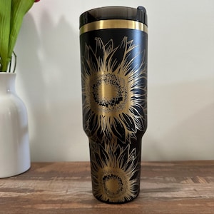 Puede incluir: Vaso t&eacute;rmico negro con un dise&ntilde;o de girasol dorado. El vaso tiene una tapa negra y una banda dorada en la parte superior. El dise&ntilde;o del girasol est&aacute; grabado en la superficie negra. Capacidad aproximada de 887 ml.