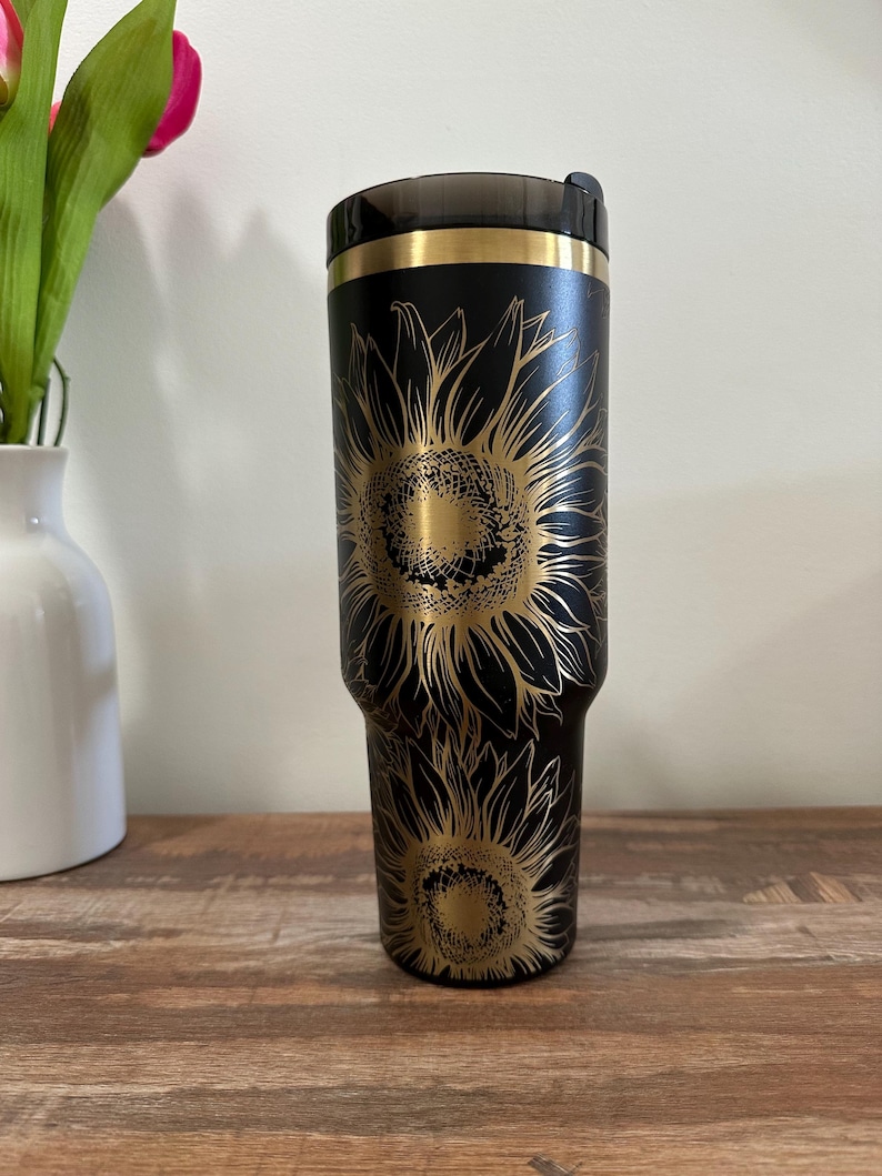 Puede incluir: Vaso t&eacute;rmico negro con un dise&ntilde;o de girasol dorado. El vaso tiene una tapa negra y una banda dorada en la parte superior. El dise&ntilde;o del girasol est&aacute; grabado en la superficie negra. Capacidad aproximada de 887 ml.