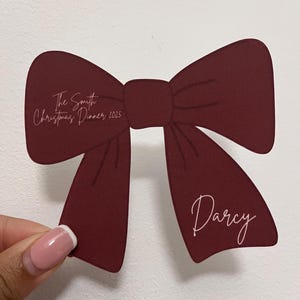 May include: A burgundy bow-shaped place card with the text "The Smith Christmas Dinner 2025" and the name "Darcy" in a cursive font. The place card is held by a hand with a white-tipped manicure.