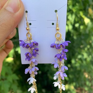 May include: A pair of dangling earrings featuring cascading purple and white flower clusters. The earrings have gold-colored hooks and chains, with clear crystal beads adding sparkle. The floral design is delicate and eye-catching.