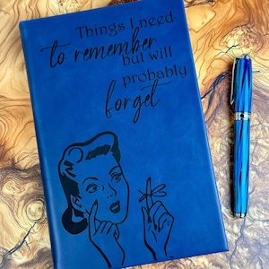 May include: A blue journal with the text "Things I need to remember but will probably forget" and a retro illustration. A blue and silver pen rests beside the journal. The journal has a retro illustration of a woman.