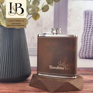 May include: A brown leather-wrapped stainless steel flask featuring a silver screw-top cap and white stitching along the edges. The front of the flask is engraved with the word Sunshine and a stylized sun graphic. The flask has a rectangular shape.