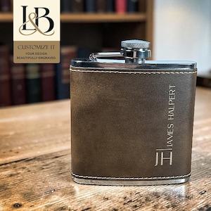 May include: A rectangular stainless steel flask wrapped in brown leather with white stitching. The leather features the engraved text "JAMES HALPERT" and the initials "JH". The flask has a silver-colored metal screw-top cap at the center.