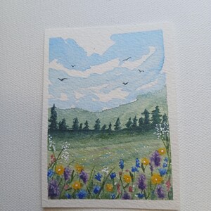 May include: A watercolor painting on textured paper depicting a landscape with a field of colorful wildflowers in the foreground, a line of dark green pine trees in the middle ground, and a light blue sky with white clouds and birds flying above.