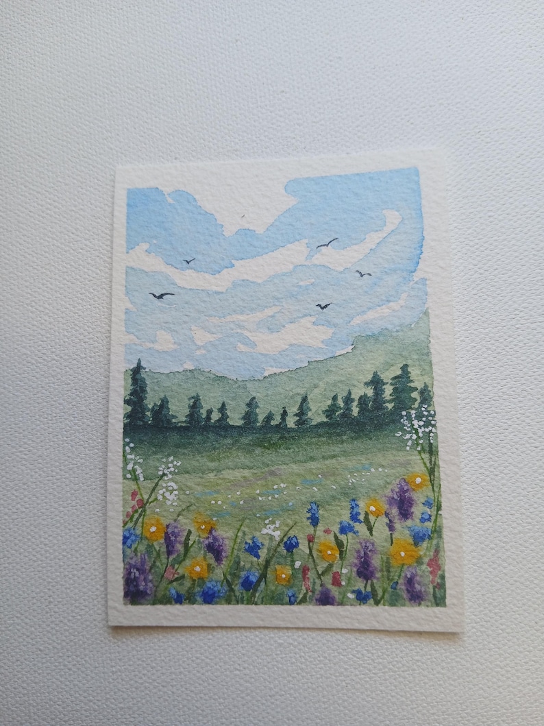 May include: A watercolor painting on textured paper depicting a landscape with a field of colorful wildflowers in the foreground, a line of dark green pine trees in the middle ground, and a light blue sky with white clouds and birds flying above.