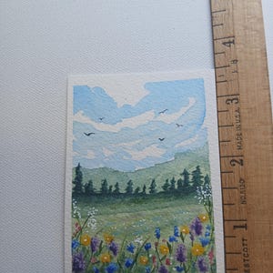May include: A rectangular watercolor painting of a landscape featuring a field of yellow, purple, and blue wildflowers in the foreground, a dark green forest, and a light blue sky with clouds and birds, next to a ruler measuring 5 inches.