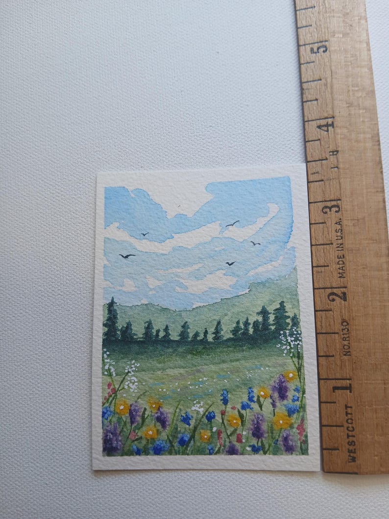 May include: A rectangular watercolor painting of a landscape featuring a field of yellow, purple, and blue wildflowers in the foreground, a dark green forest, and a light blue sky with clouds and birds, next to a ruler measuring 5 inches.