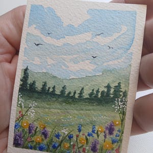 May include: A small rectangular watercolor painting on paper depicting a landscape with a field of colorful wildflowers in the foreground, a line of dark green pine trees in the middle ground, and a light blue sky with white clouds and birds.