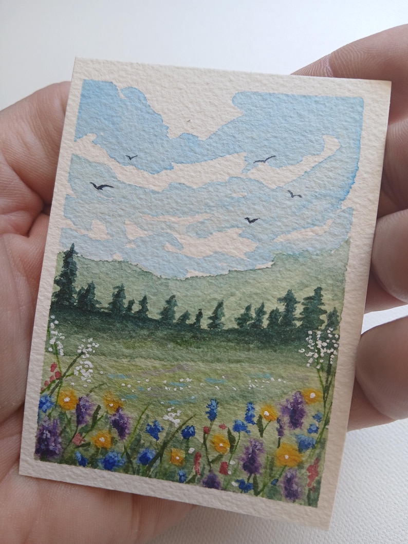 May include: A small rectangular watercolor painting on paper depicting a landscape with a field of colorful wildflowers in the foreground, a line of dark green pine trees in the middle ground, and a light blue sky with white clouds and birds.