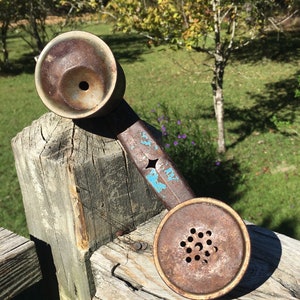 May include: A vintage telephone handset featuring a heavily rusted metal surface with patches of chipped blue paint. The device includes a circular earpiece and a perforated mouthpiece, showing significant signs of age, oxidation, and wear.