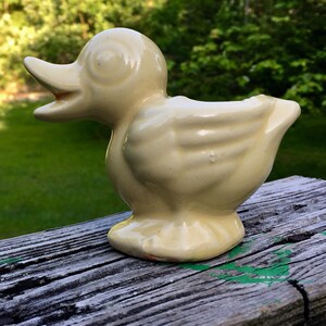 May include: A pale yellow ceramic duck figurine with a glossy finish. The duck is depicted in profile, facing left with its beak slightly open. The sculpture features molded wing details and a smooth, rounded body shape, standing on a flat base.