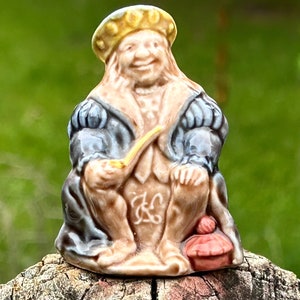 May include: A small, detailed figurine of a seated man in historical attire. The figure wears a gold hat, a blue and tan coat, and holds a pipe. The figurine has a smiling face and is set on a wooden surface. A small red hat is at the figure's feet.