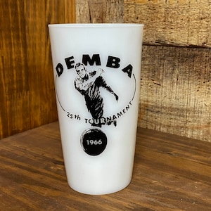 May include: A white milk glass tumbler featuring a black graphic of a person bowling. The text reads "DEMBA 25th TOURNAMENT" in an arc above the figure, with a black bowling ball at the bottom containing the year "1966" printed in white text.